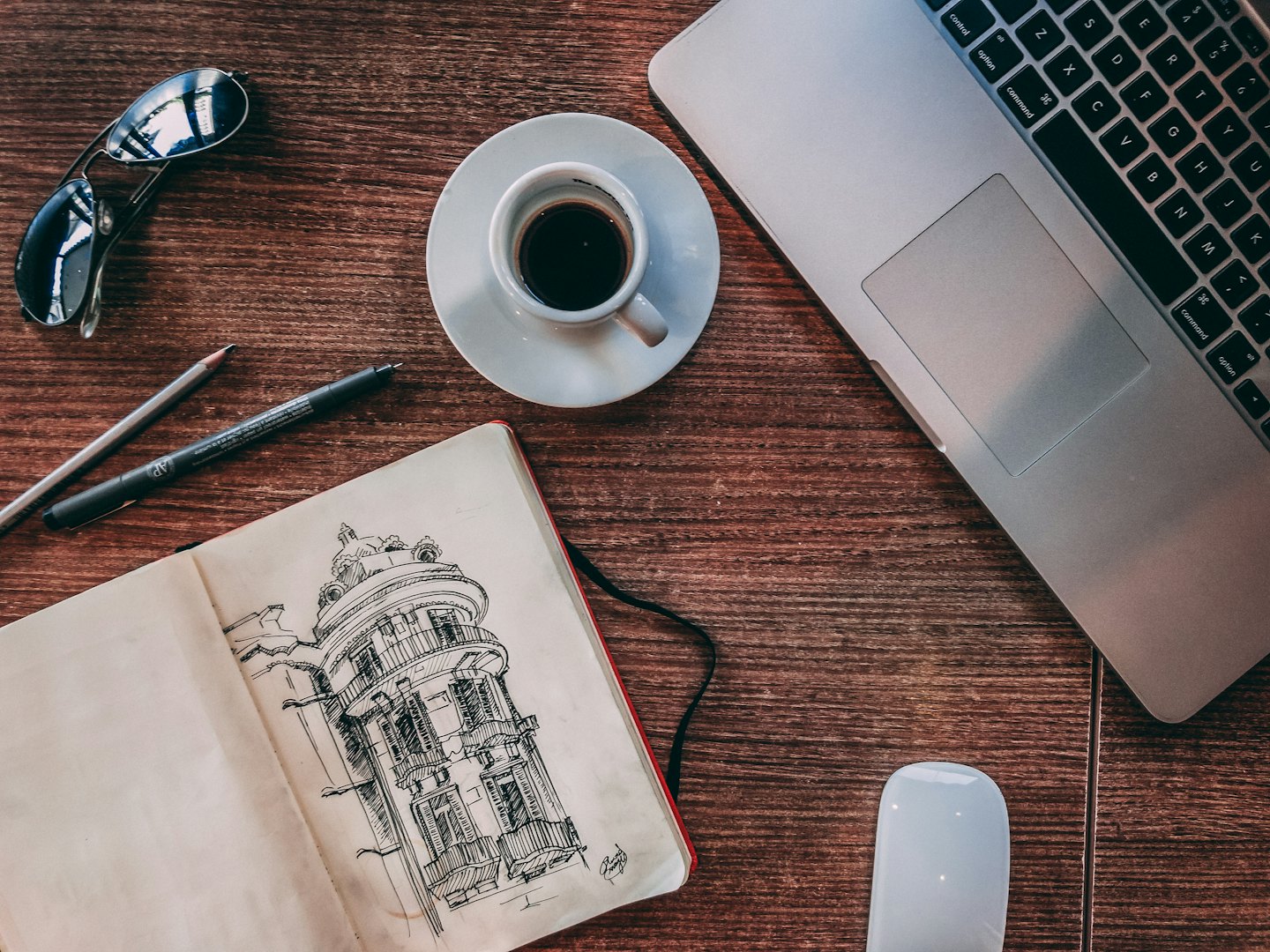 Creative workspace with sketchbook, laptop, and coffee on wooden table.